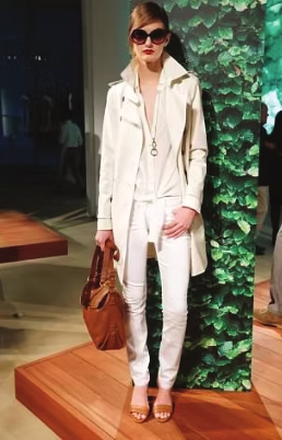 Woman in white outfit with sunglasses and brown bag, standing by greenery.