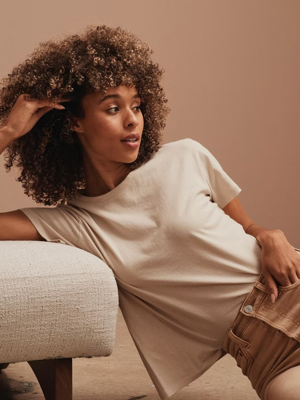 Woman with curly hair in a tan t-shirt and shorts, leaning on a beige ottoman, looking to the side.