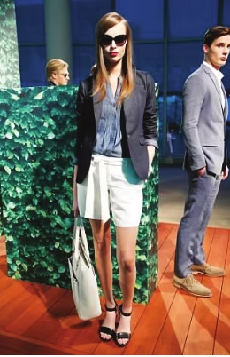 Woman in sunglasses, blazer, shorts, and sandals, holding a bag, posed next to a man in a suit.