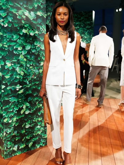 Woman in white suit and heels, holding a clutch, standing on a wooden floor in front of a green wall.