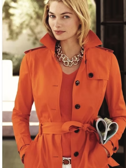 Woman in orange trench coat and silver necklace holding a magazine.