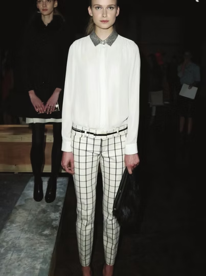 Woman in white shirt, checkered pants, and holding a black purse, posing. Another person stands in the background.