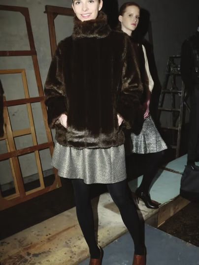 Woman in a brown fur jacket, silver skirt, and black tights. Another woman in background. Indoors.