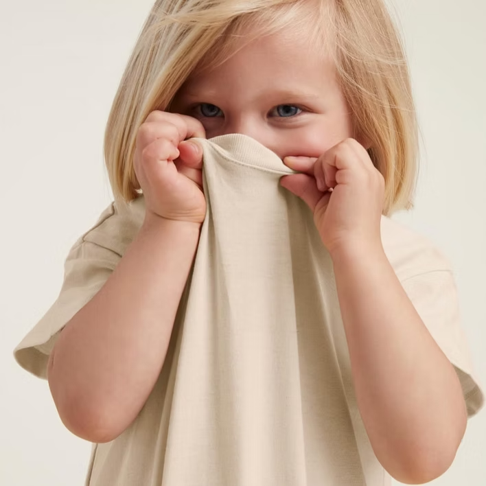 Blond child holding up the collar of a beige t-shirt, looking shyly at the camera.
