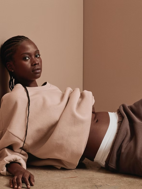 Woman in beige sweatshirt and brown pants reclines against a tan wall, looking towards camera.