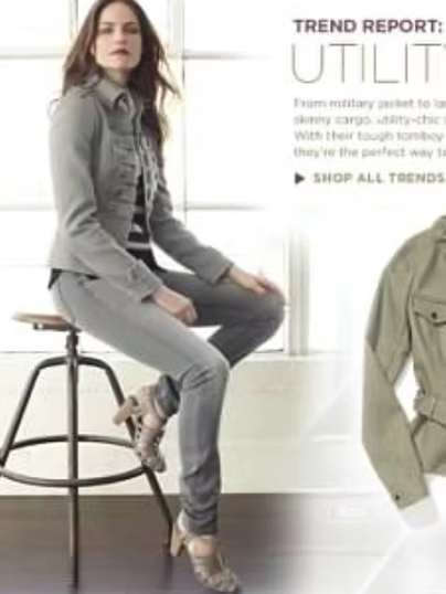 Woman in grey utility jacket and jeans sits on stool, looking towards the viewer.