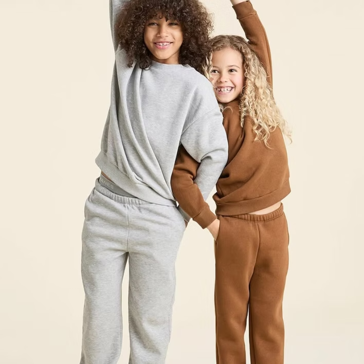 Two children in matching sweatsuits, one gray, one brown, smiling, arms raised.