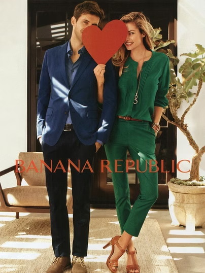 Couple holding a heart in front of faces; man in blue jacket, woman in green jumpsuit, inside a Banana Republic.