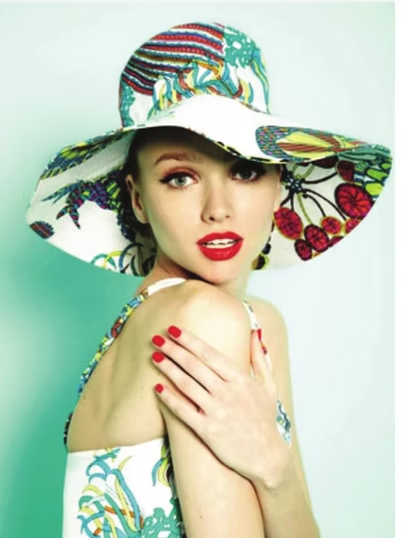 Woman in floral hat and dress, red lipstick and nails, light blue background.
