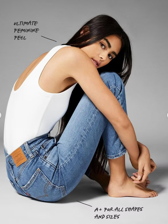 Woman in white tank top and blue jeans, seated with legs crossed, looking at the viewer.