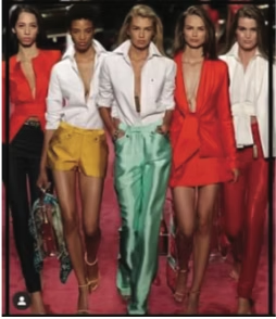 Five models in bright outfits walk a runway. A mix of pants, shorts, and blazers in red, white, yellow, and green.