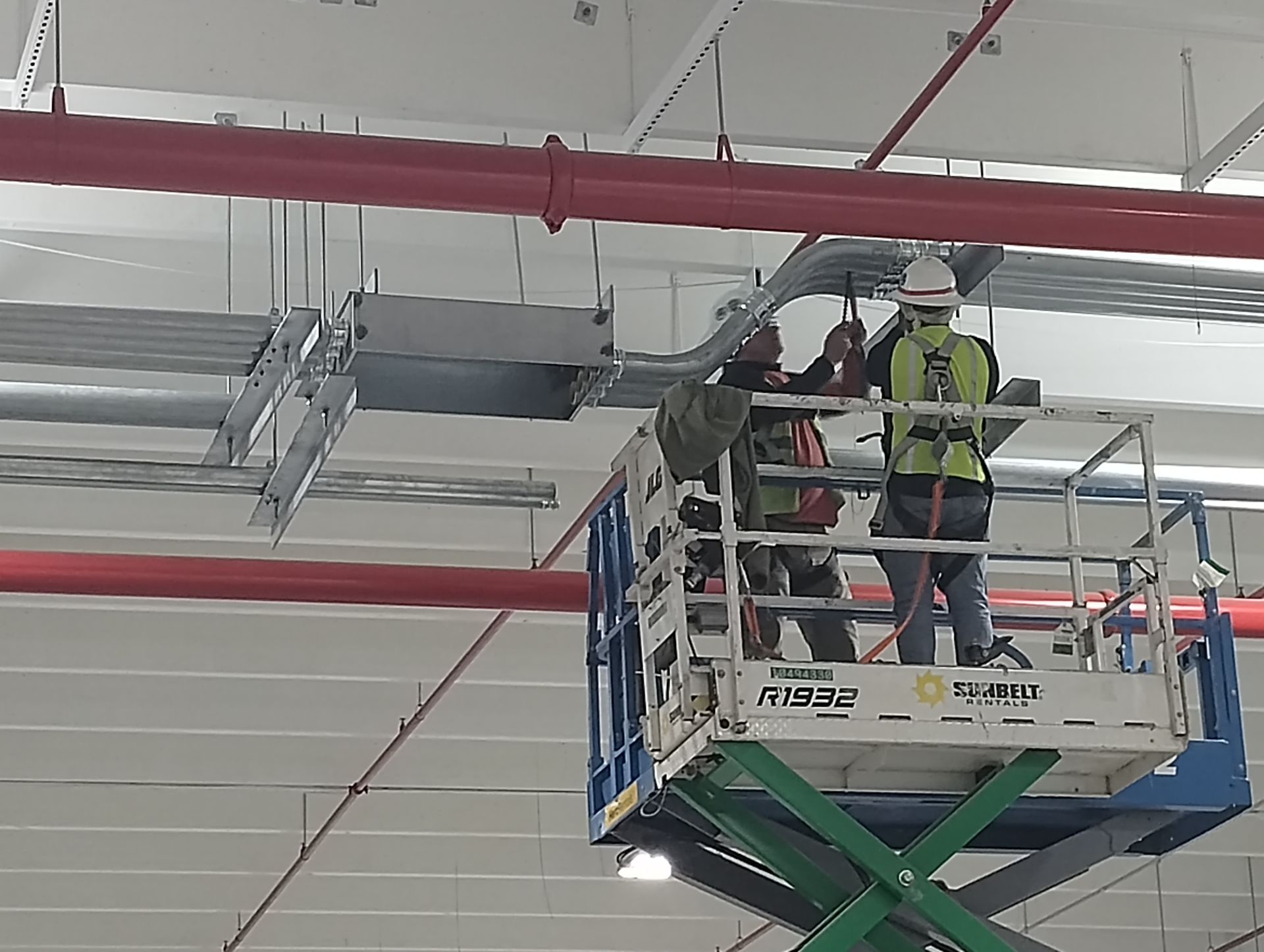 Two workers on a lift installing ductwork near a red pipe in a large, industrial space.