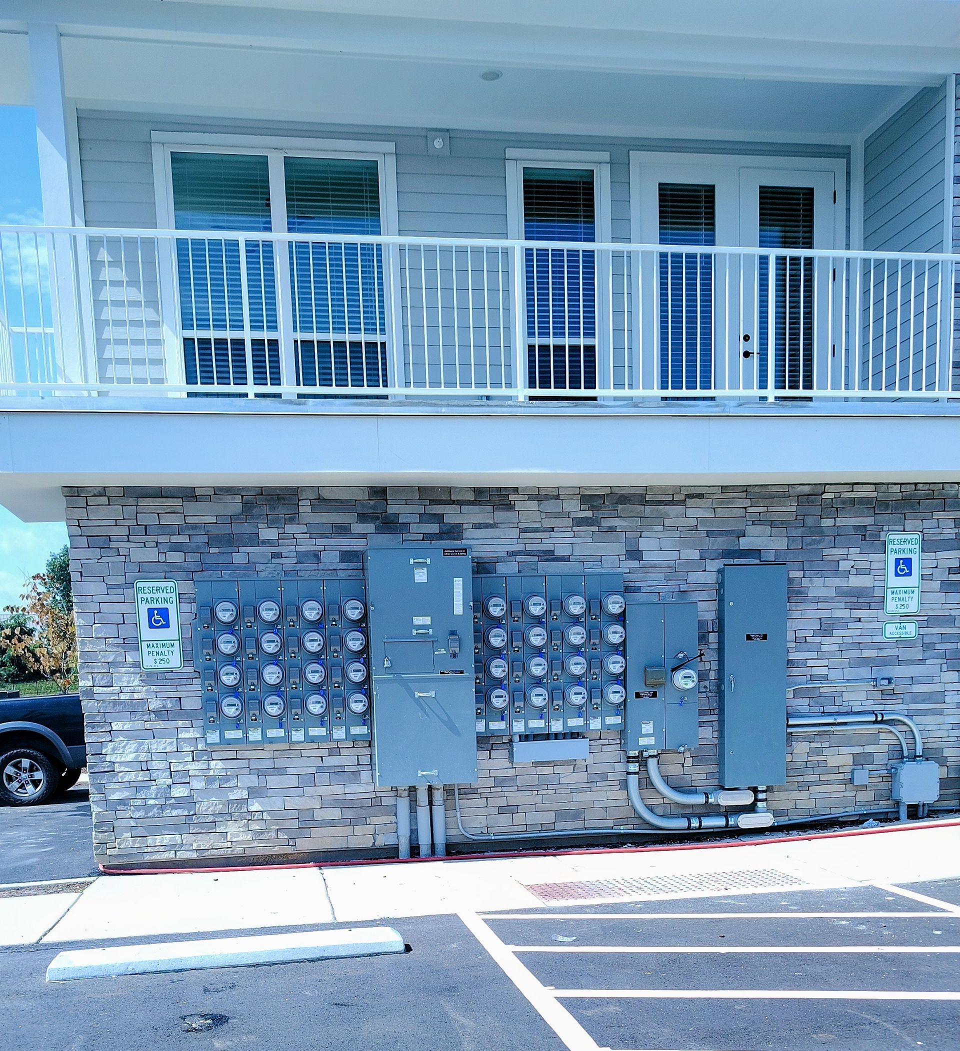 Gray apartment building exterior with utility boxes on a stone wall below a balcony.