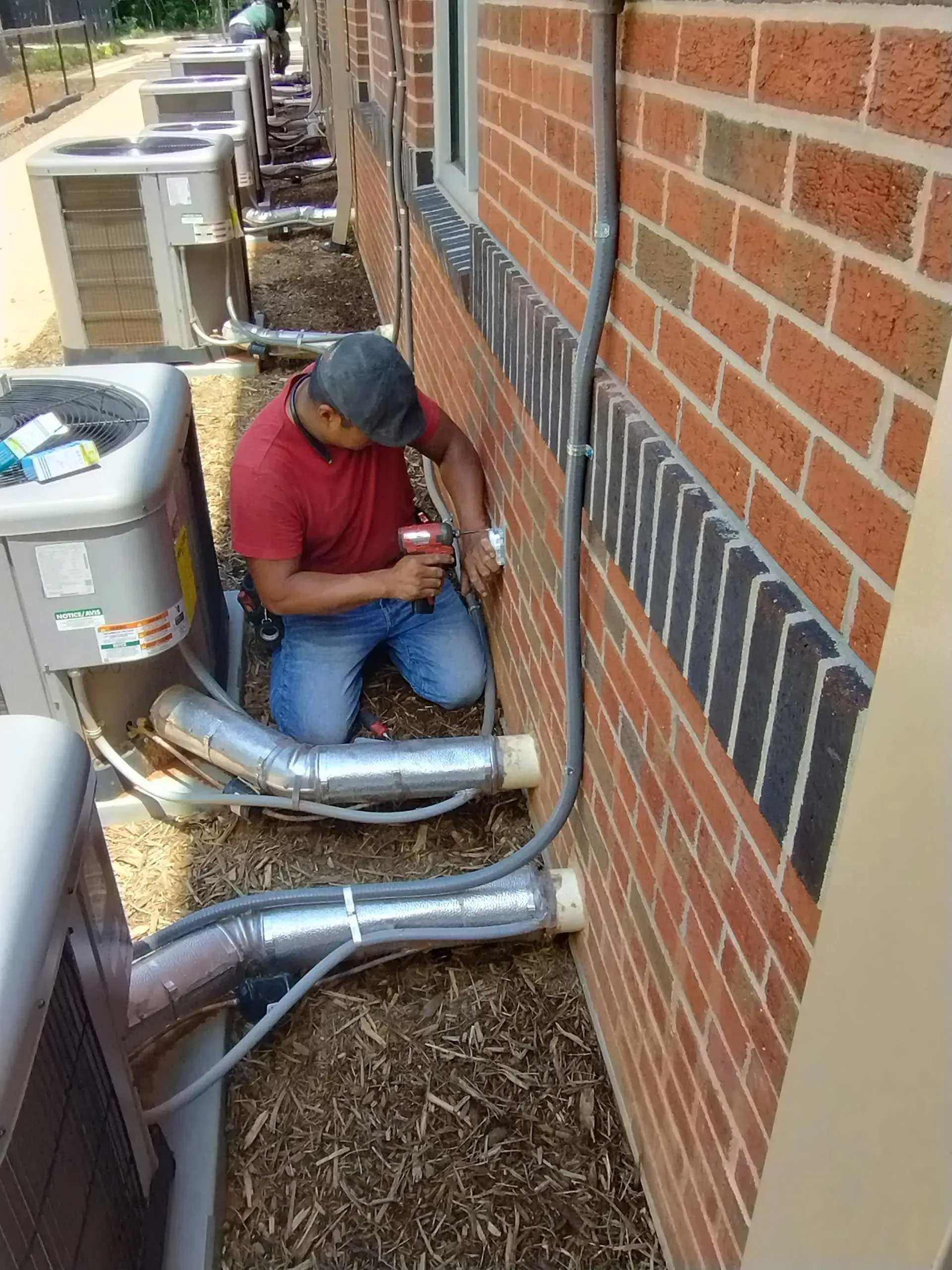 Man in jeans and red shirt working on electrical conduit near brick building and AC units.