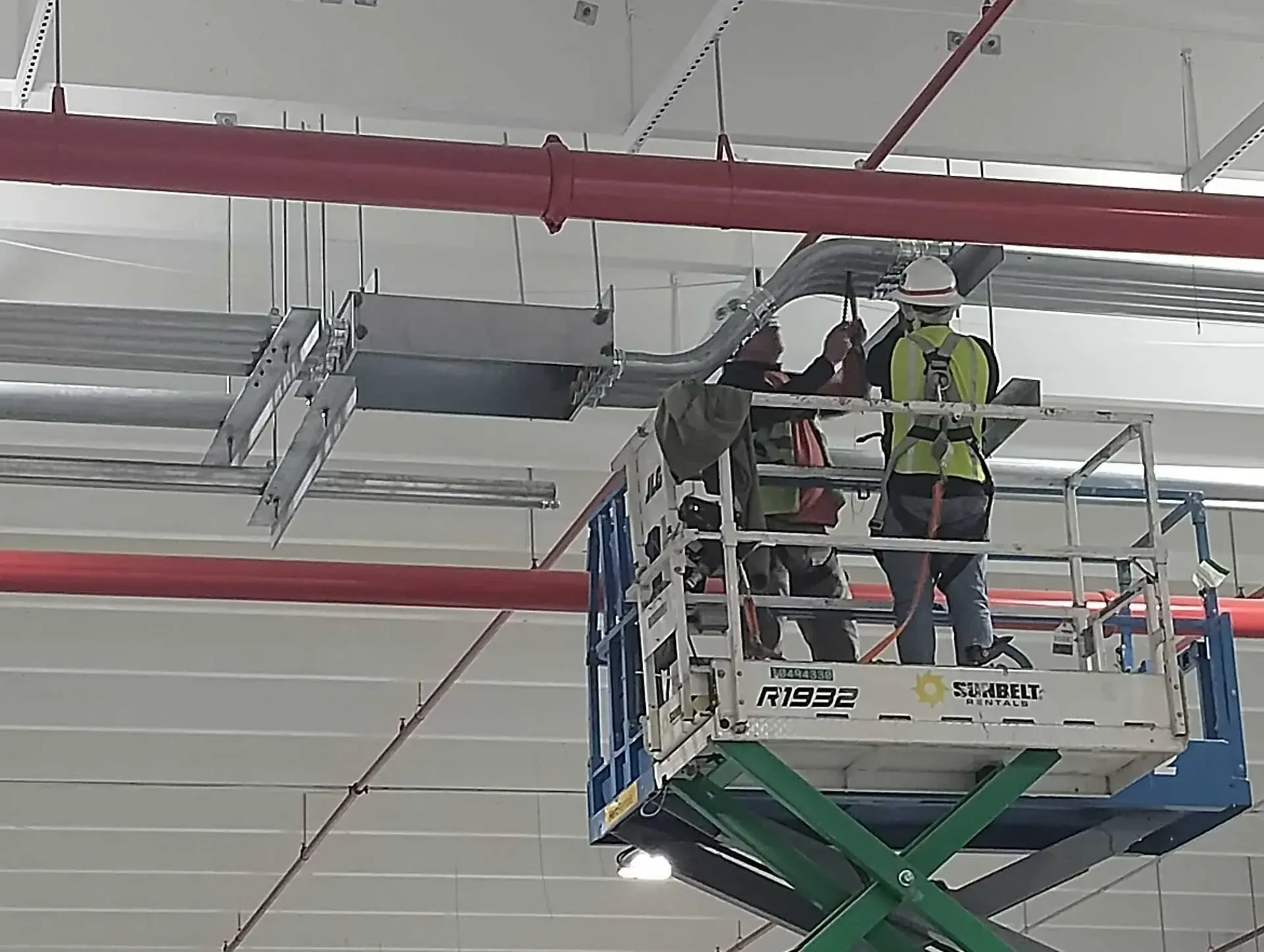 Two construction workers on a lift installing cables in a warehouse, near red pipes.