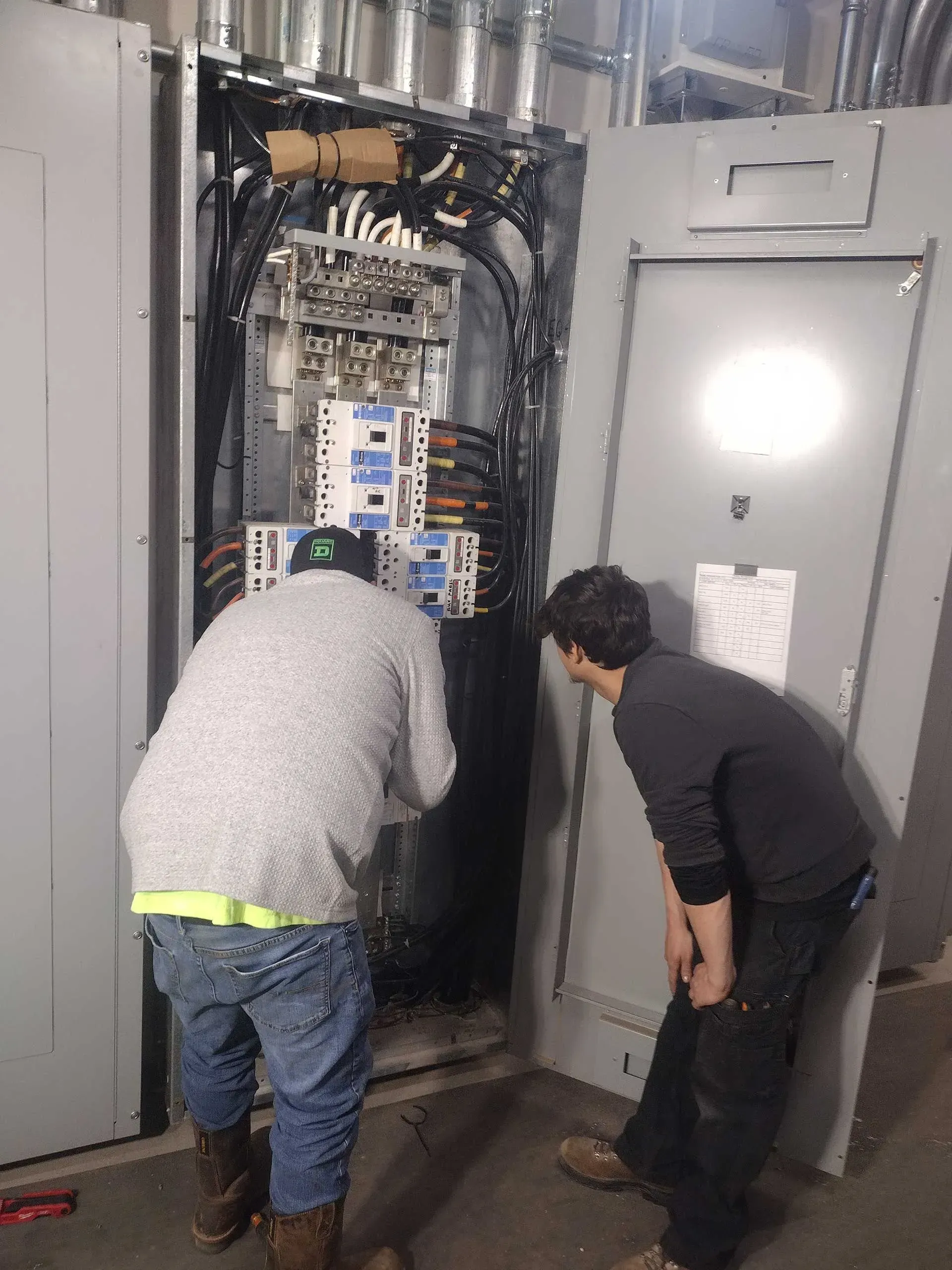 Two people inspecting an electrical panel. One wears a white sweater and jeans, the other a black shirt. The panel is open.