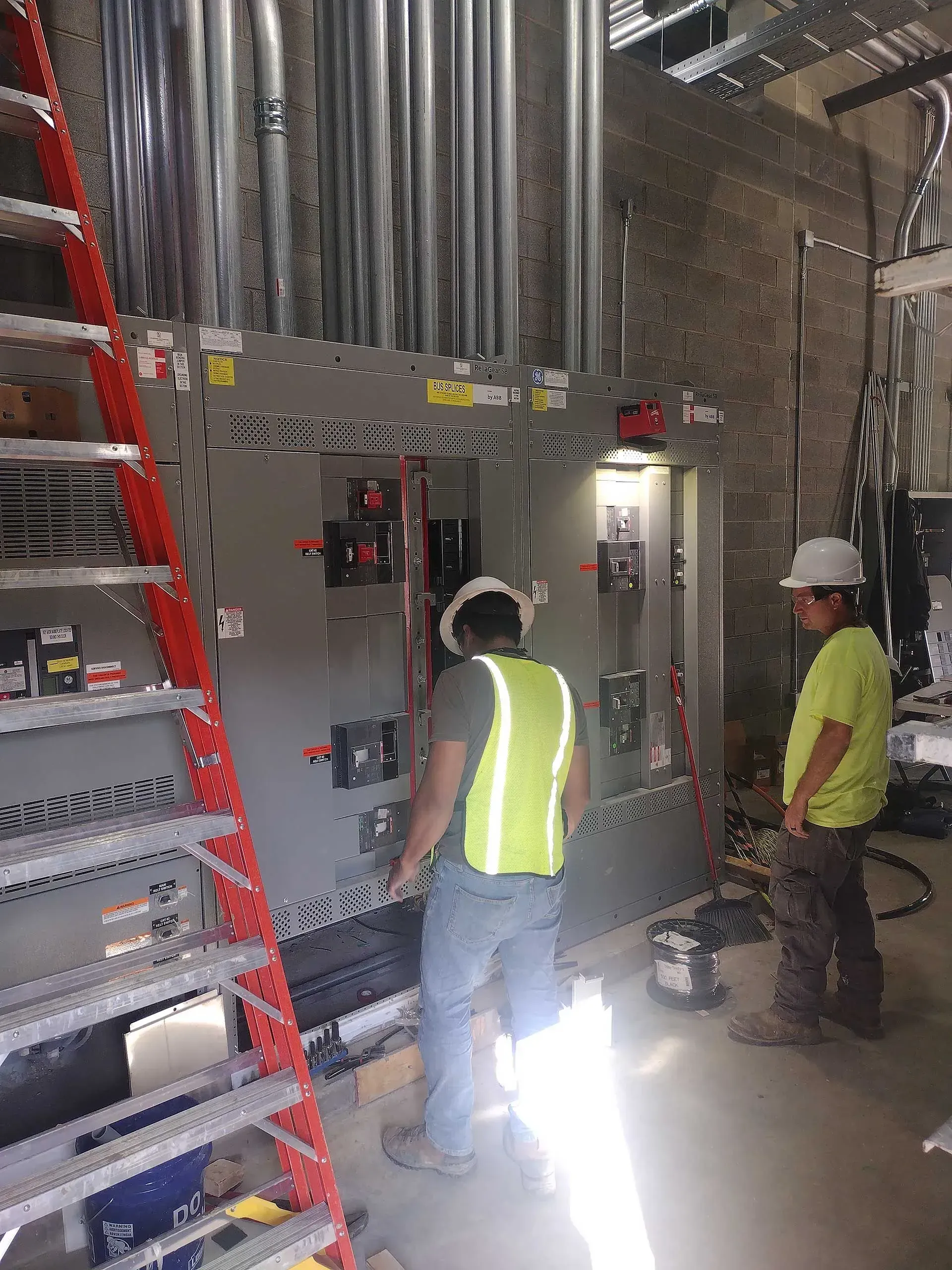 Two workers in safety vests working on electrical panel. Conduit pipes run above it, ladder on the left.