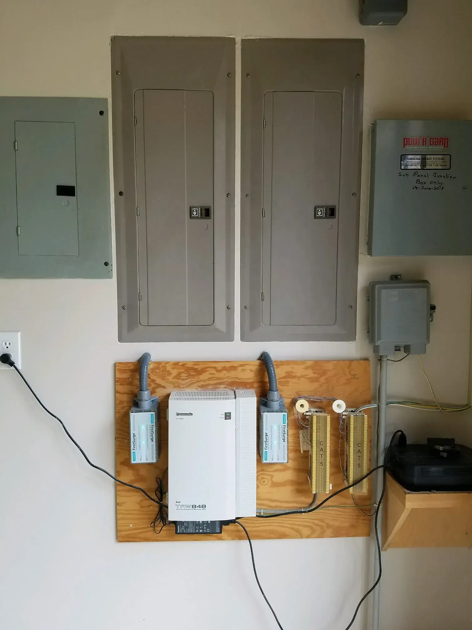 Electrical panels, conduit, and wiring on a beige wall, including a telephone, and a wooden board with electronic equipment.