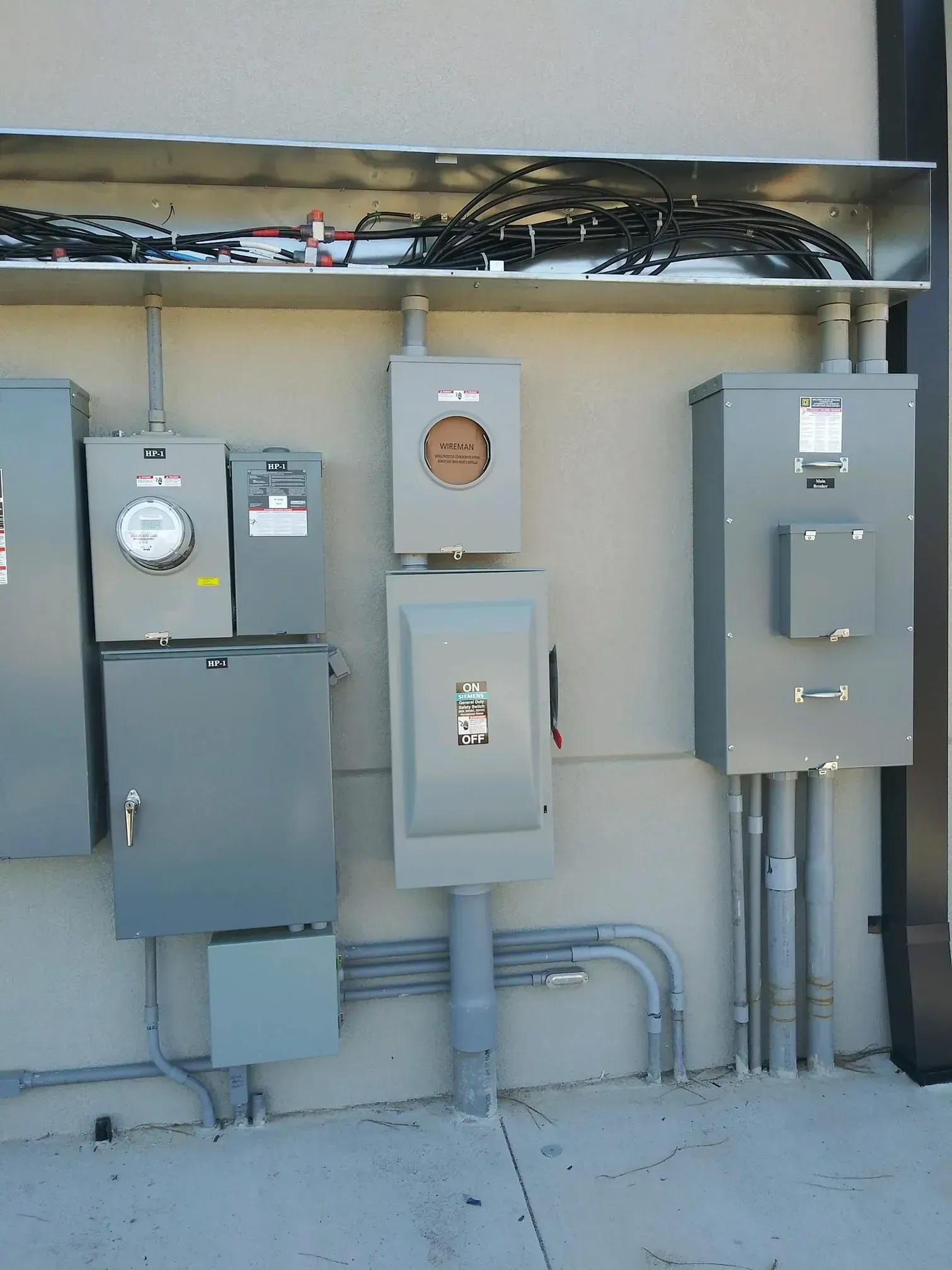 Gray electrical boxes and conduit mounted on a light tan building exterior. Wiring runs along a shelf above.
