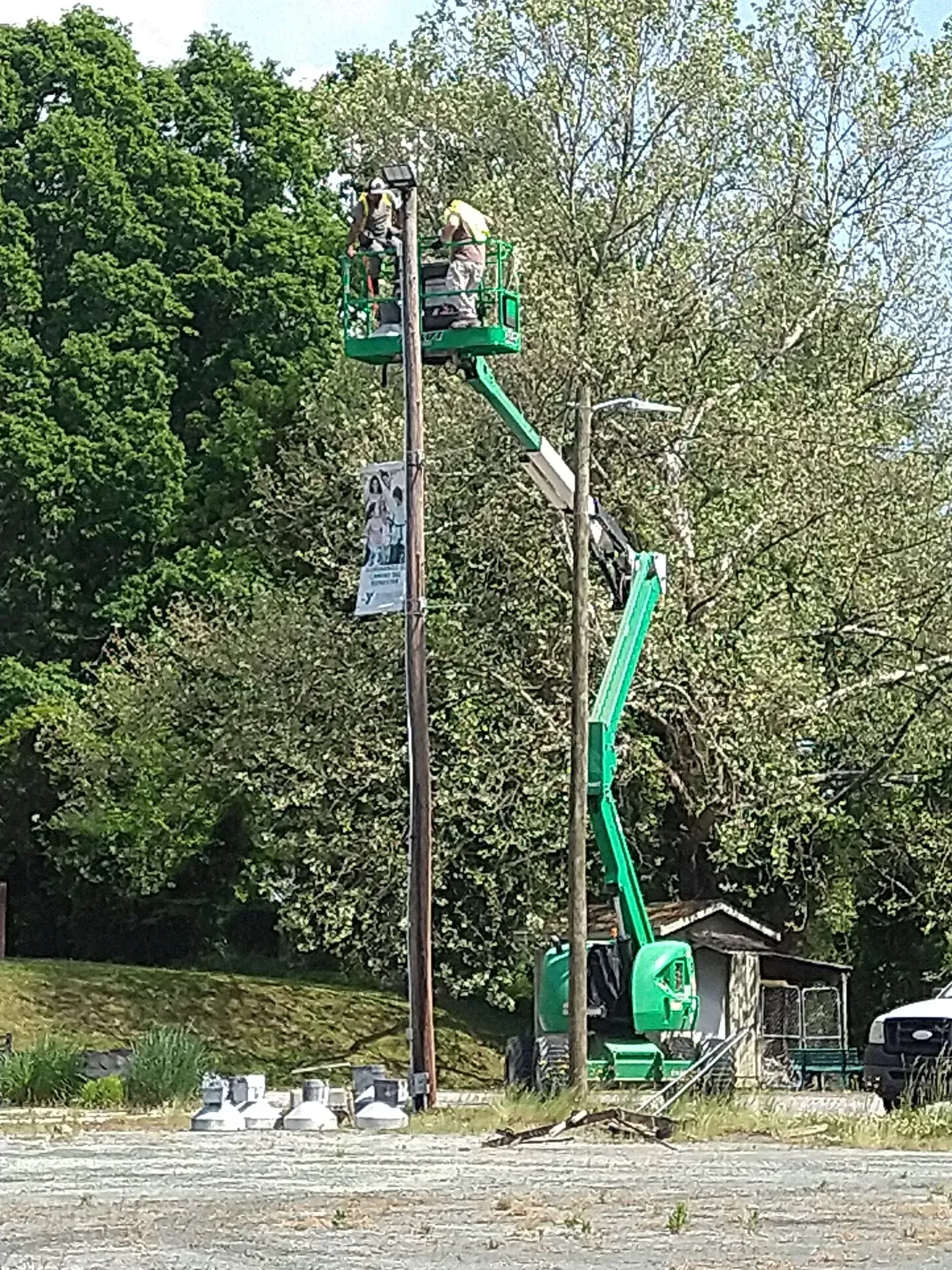 Workers on a lift are repairing utility lines on a pole. Green lift, trees, a small building, and a parked vehicle are in view.