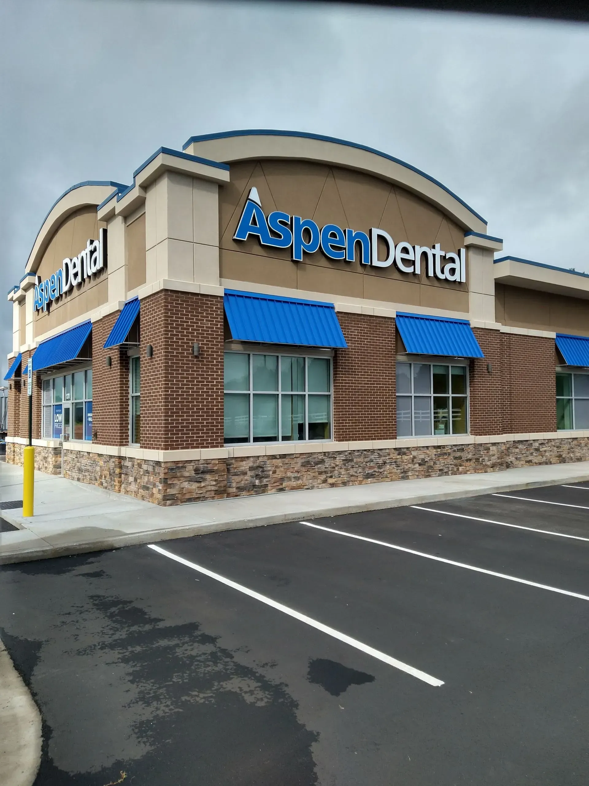 Aspen Dental building with blue awnings and signage, beige and brick facade, and a cloudy sky.
