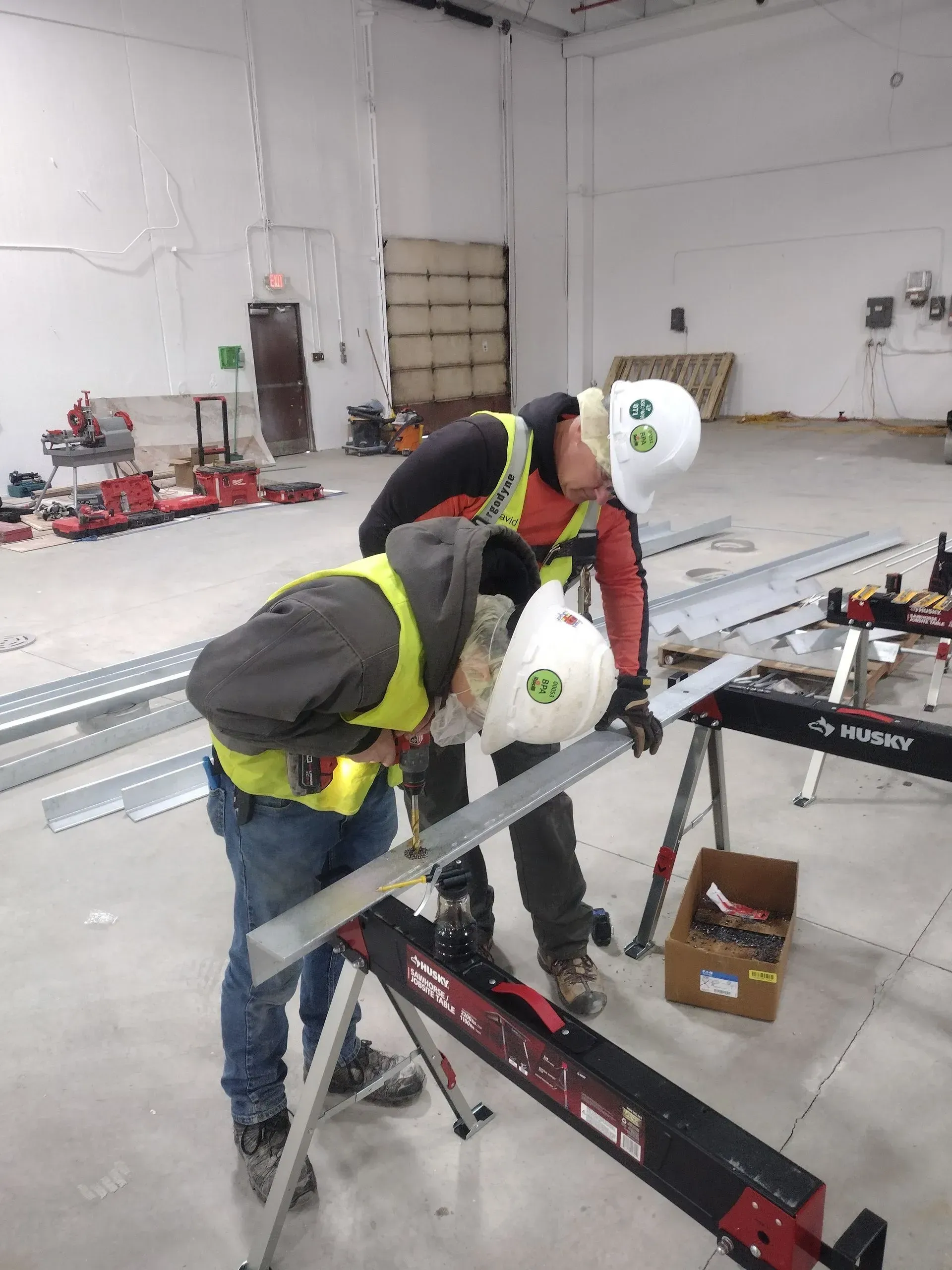Two construction workers wearing safety vests and hard hats, cutting metal with a saw at a worksite.