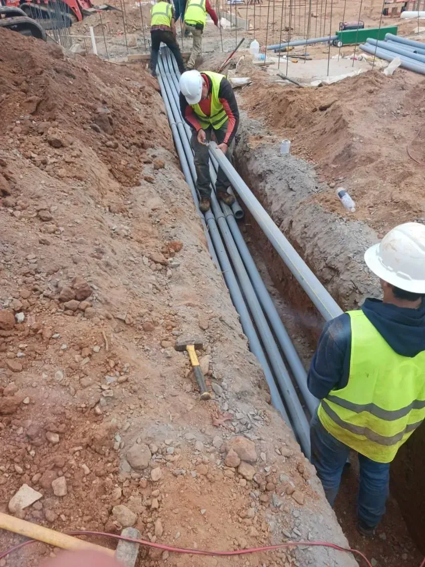 Construction workers installing gray conduit pipes in a trench at a building site.