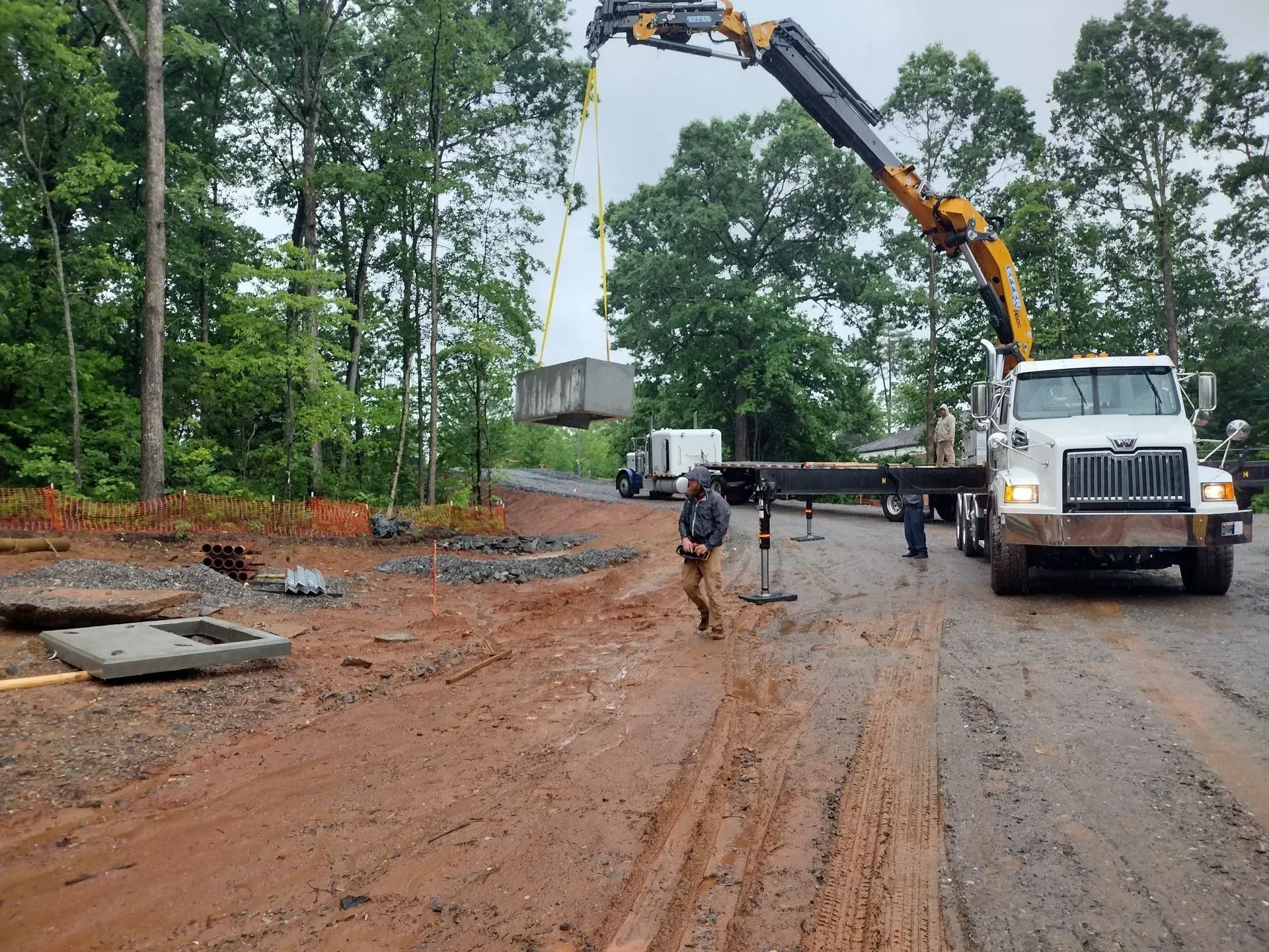 Crane lifting a concrete structure on a muddy road, workers nearby; trees in background.