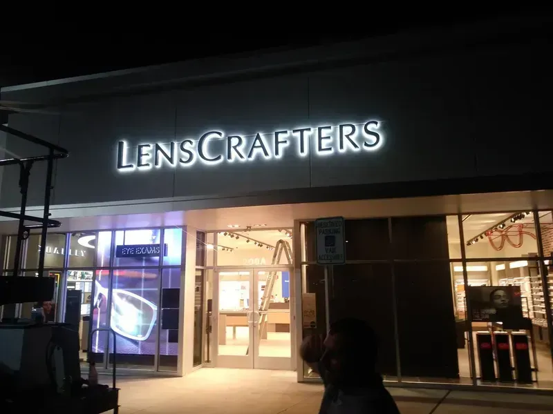 LensCrafters store front at night, illuminated sign, glass windows, and a person in the lower right.