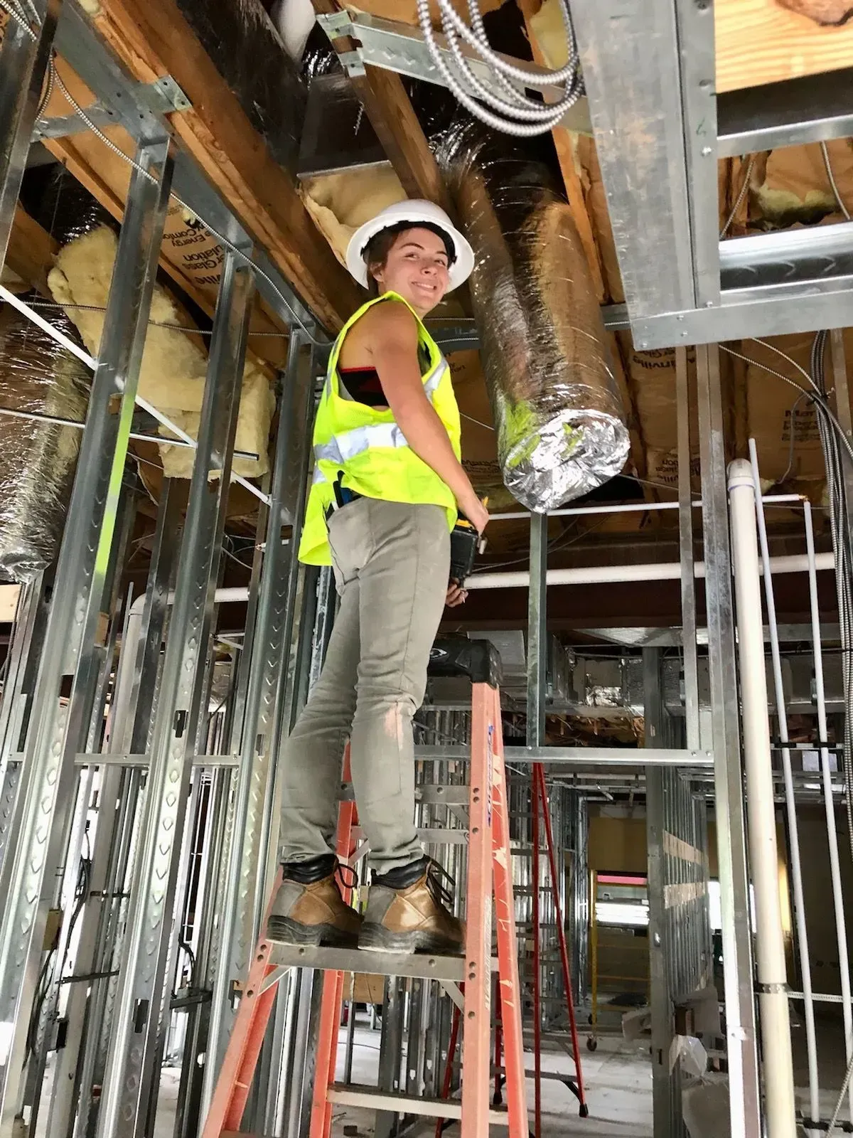 Woman in hard hat and safety vest on ladder, working on ceiling in construction site.