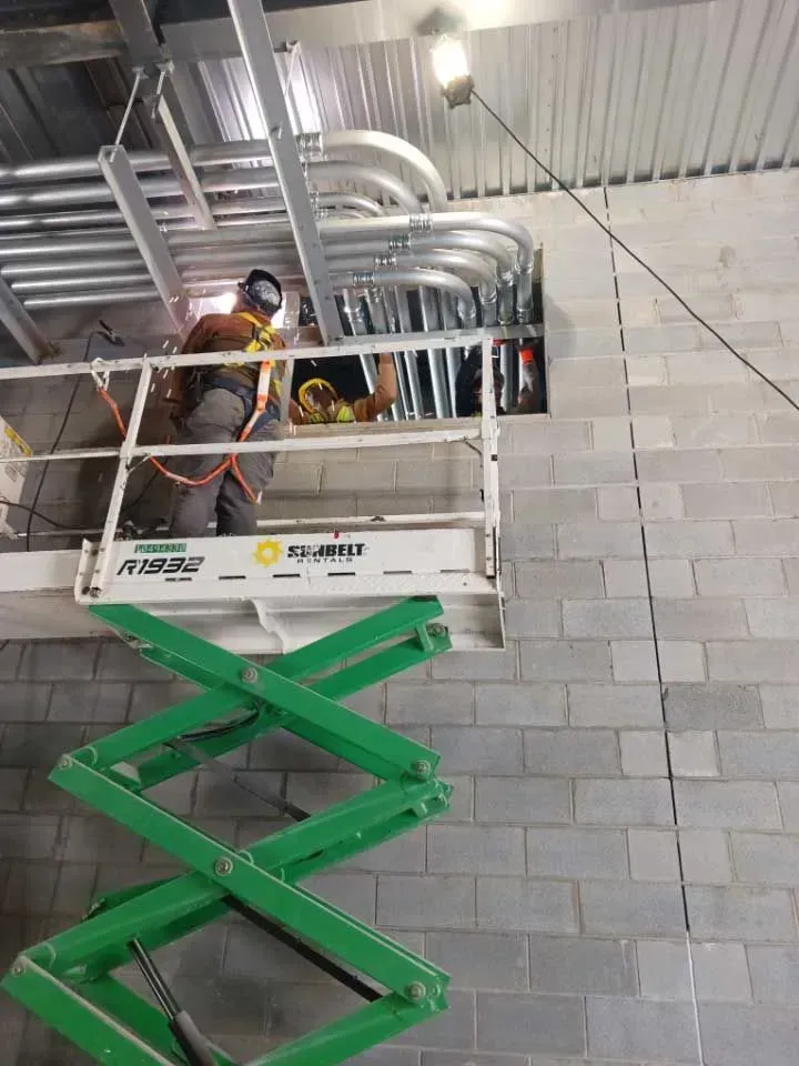 Construction workers on a lift install conduit in a wall, wearing safety gear. Inside a building with grey cinder block walls.