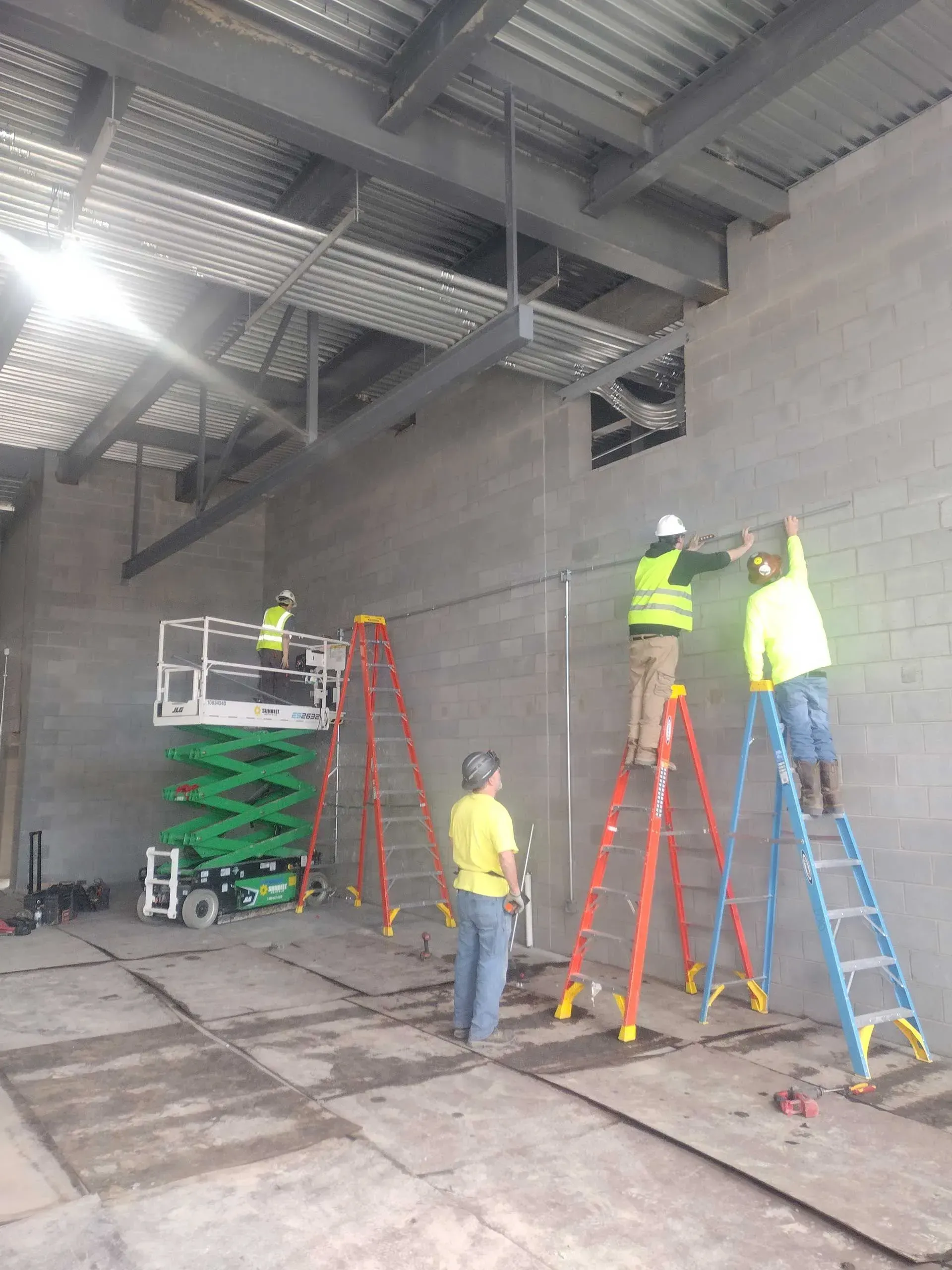 Construction workers on ladders and lift, wiring interior of building with concrete walls.