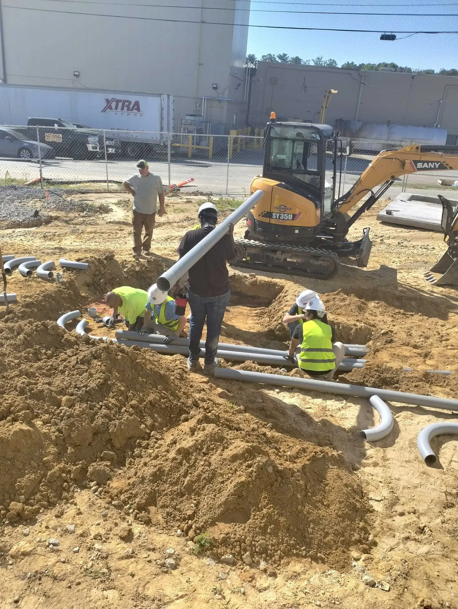 Construction workers installing pipes in a sandy area next to a small excavator and building.