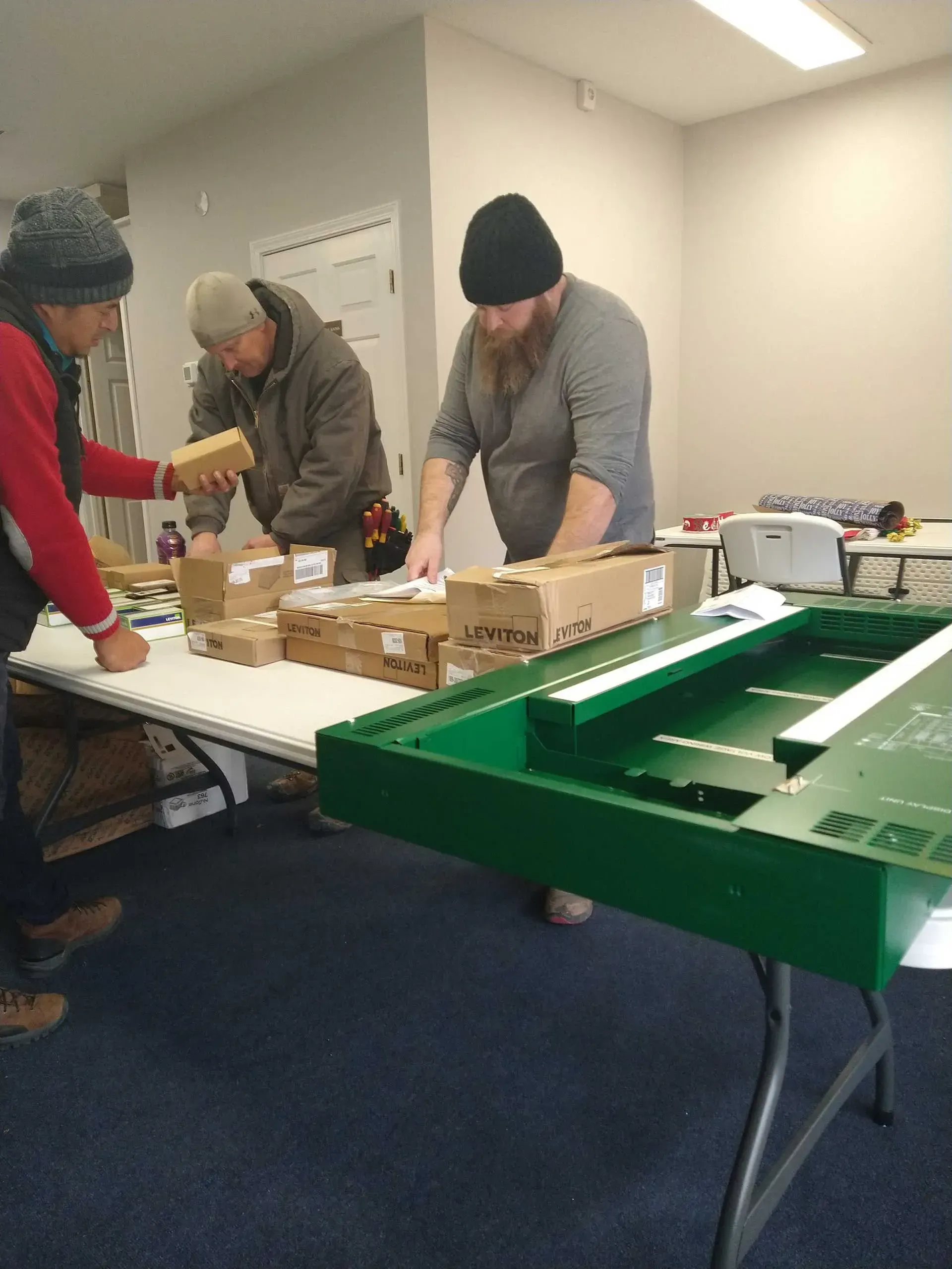 Three people assembling items at a table, with boxes and a green frame. Inside a room.