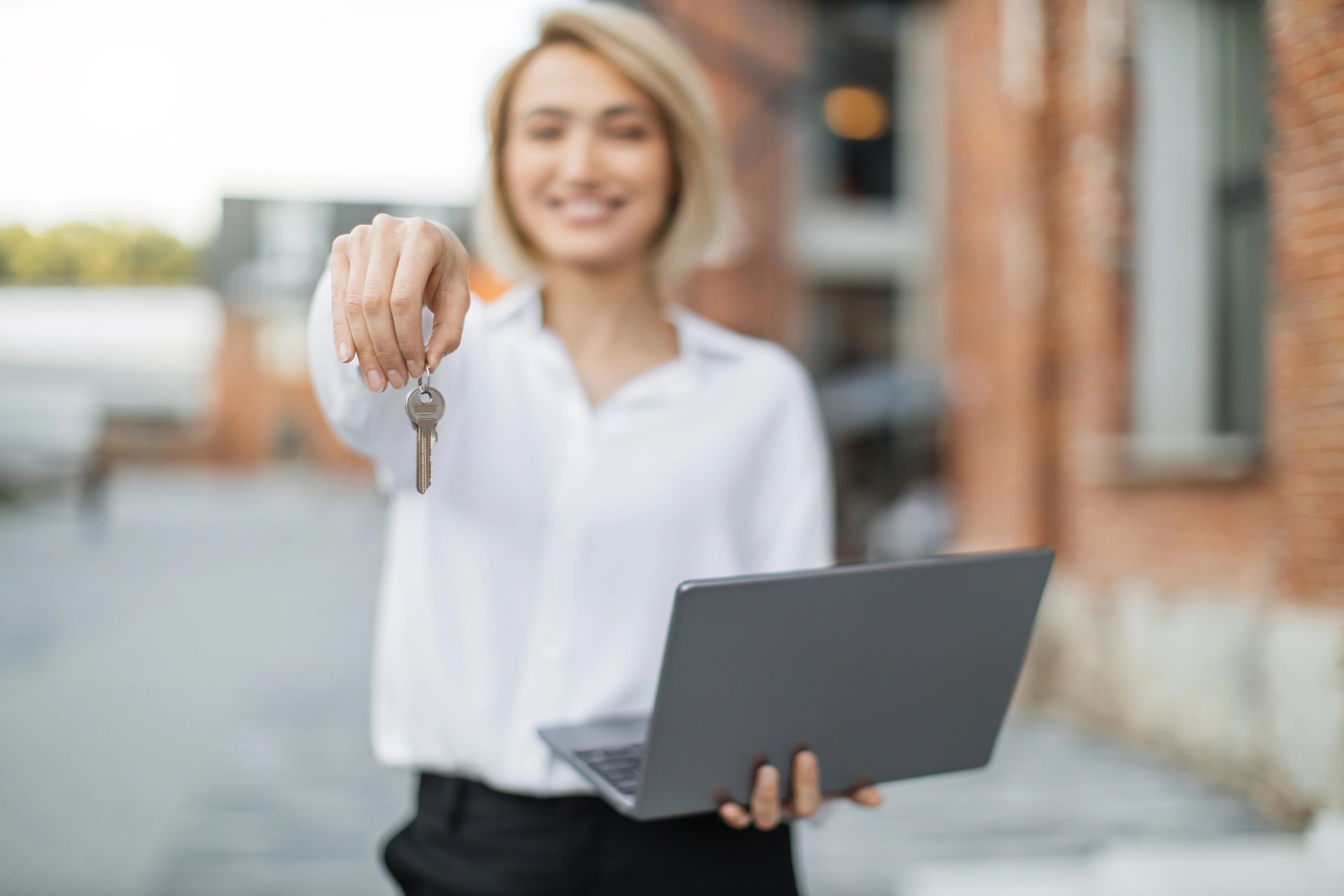Woman holding keys and laptop, smiling, in front of a brick building.