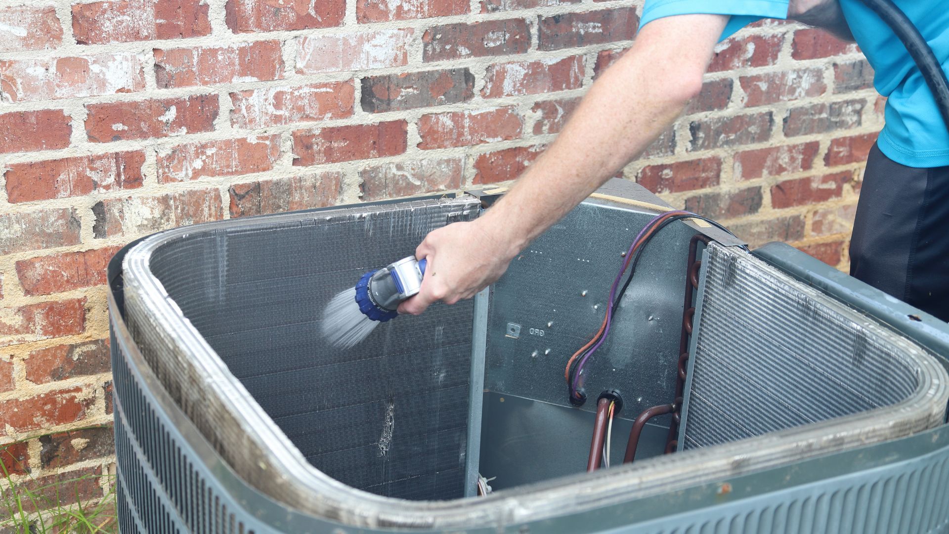 Person spraying an outdoor AC unit's metal fins with a hose near a brick wall.
