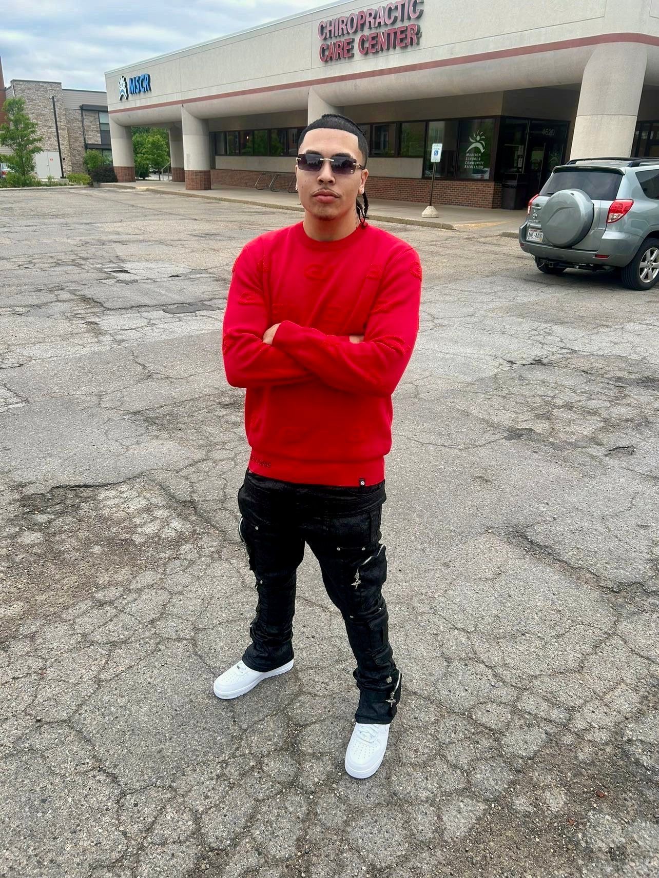 Man in red sweater and black jeans standing in a parking lot, arms crossed.