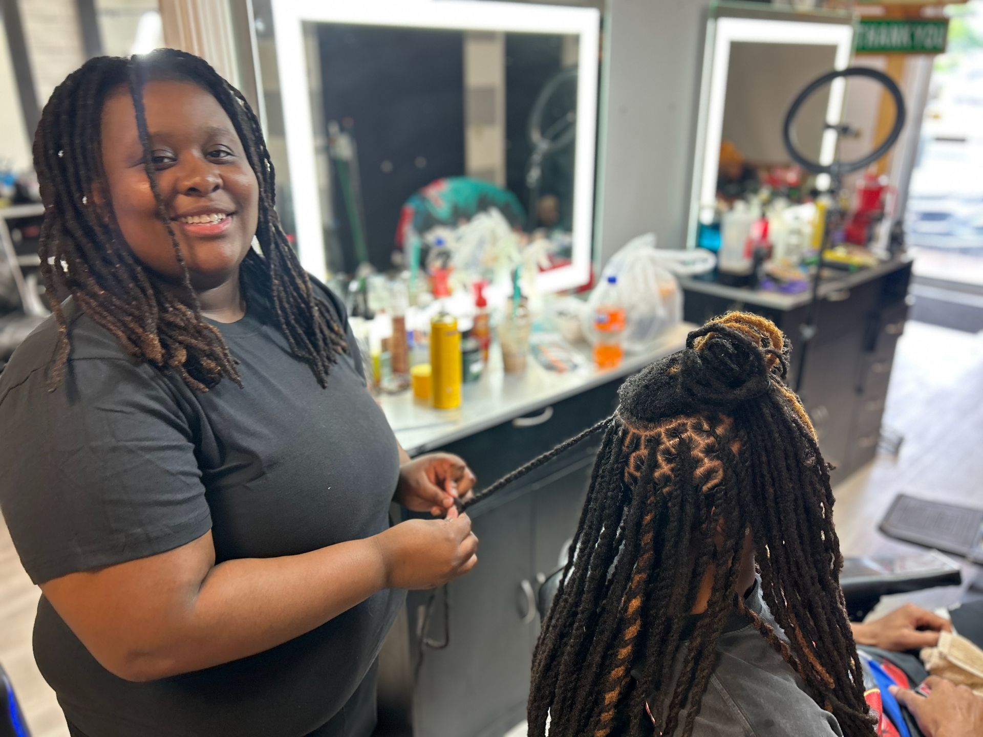 Woman with dreadlocks braiding another woman's hair in a hair salon, smiling.