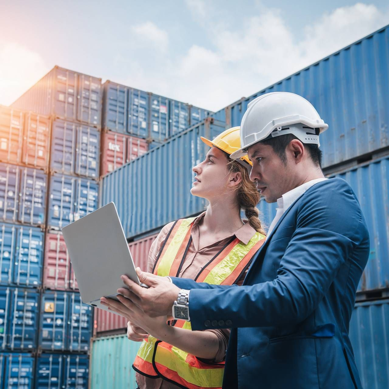 Twee professionals met veiligheidshelmen bekijken informatie op een laptop voor een stapel zeecontainers.