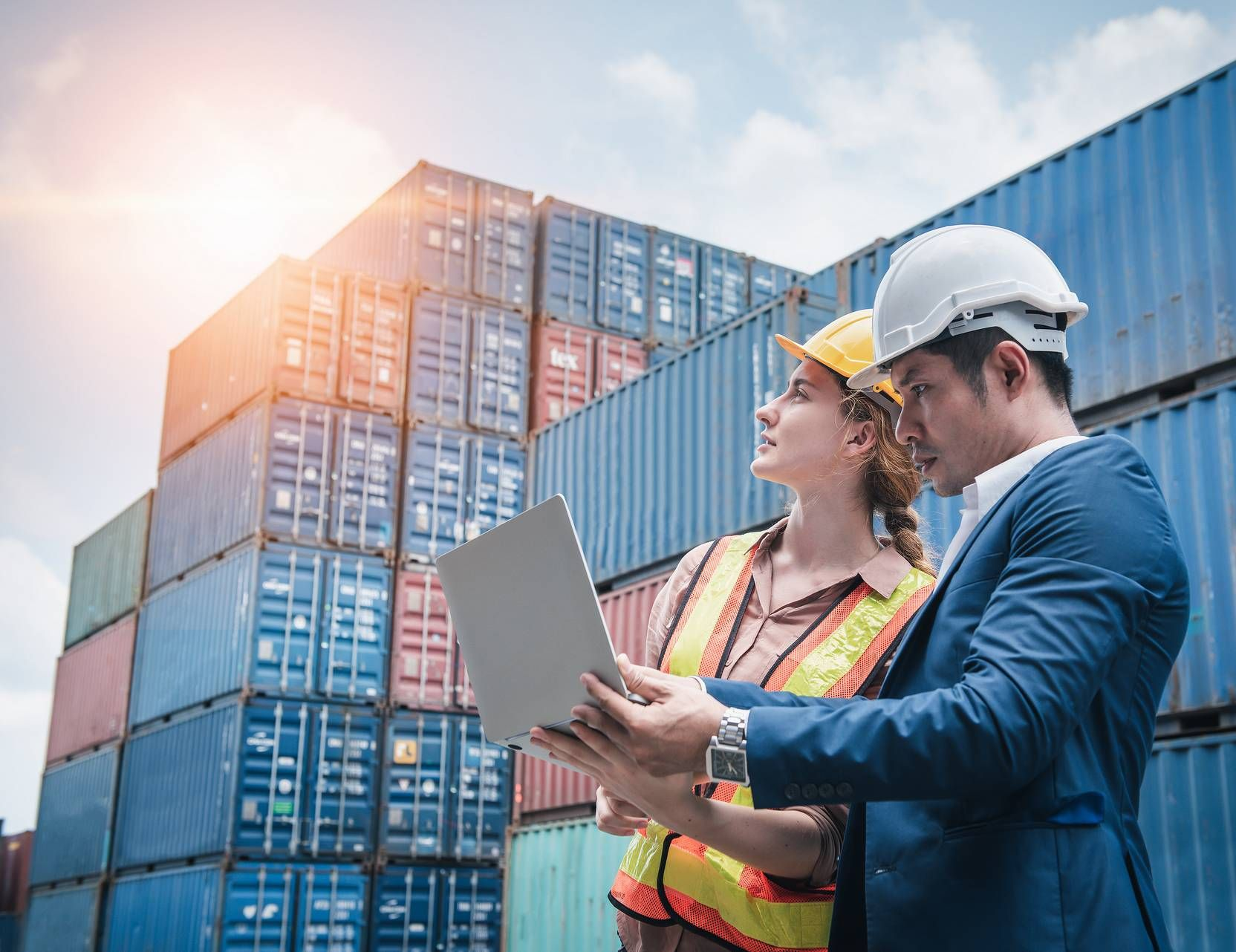 Twee professionals met veiligheidshelmen bekijken informatie op een laptop voor een stapel zeecontainers.