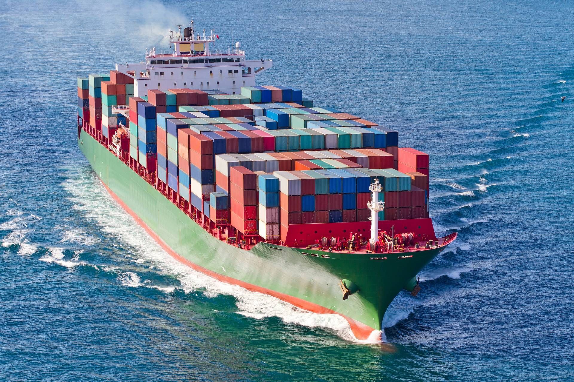 Een groot, veelkleurig containerschip vaart door het blauwe oceaanwater en laat een wit spoor achter zich.