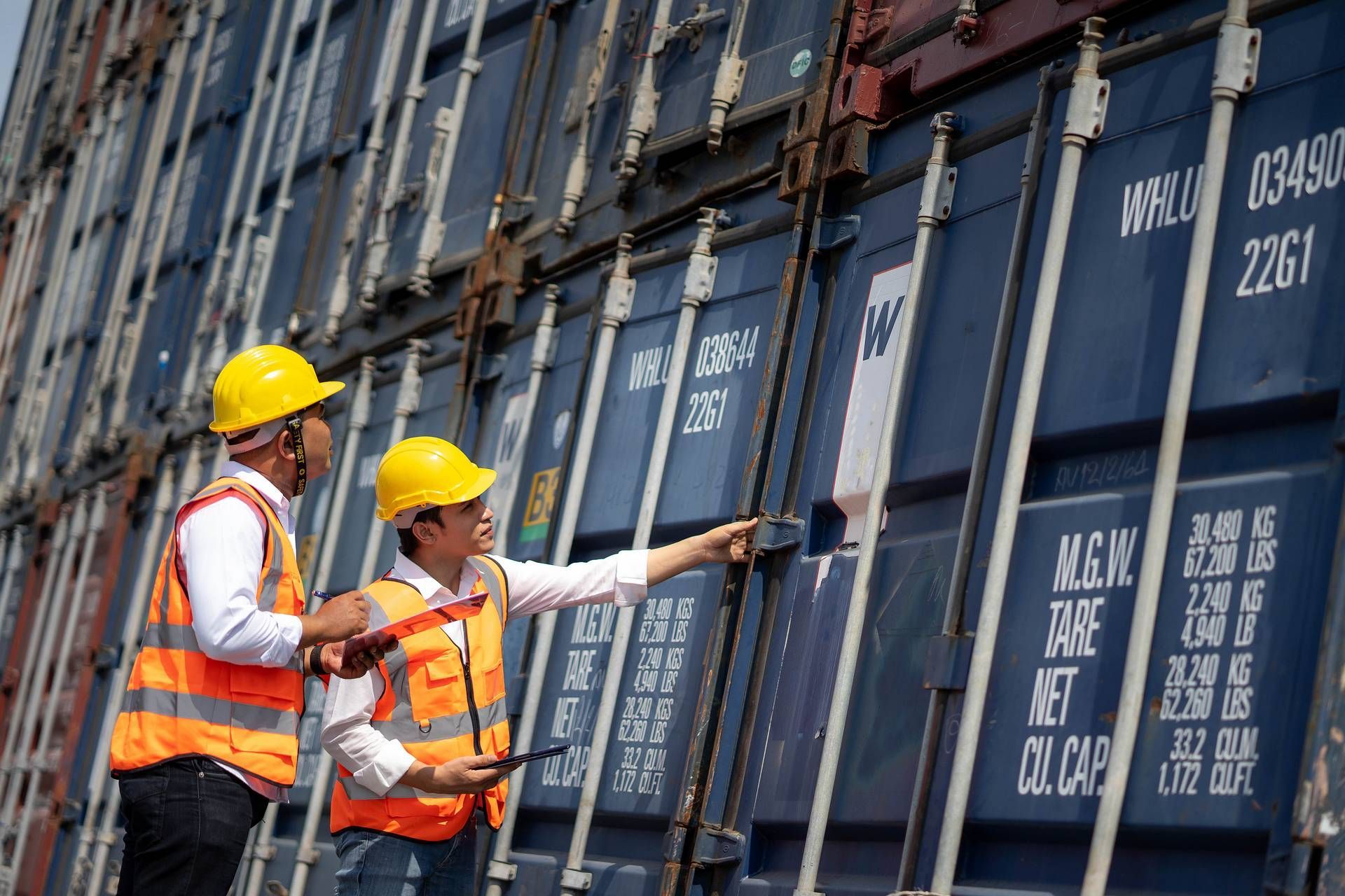 Twee werknemers met veiligheidshelmen en -vesten inspecteren zeecontainers in een havenfaciliteit.