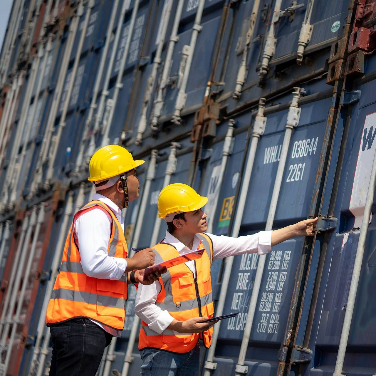 Twee werknemers in reflecterende hesjes en veiligheidshelmen inspecteren zeecontainers in een haven.