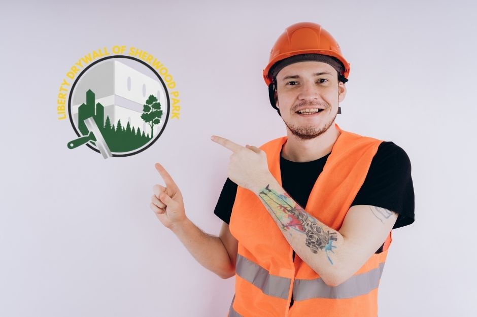 A drywall handyman smiling while wearing an orange safety vest and hard hat, pointing with both hands toward an empty light-colored wall, with a visible tattoo on his forearm and a plain indoor background in Sherwood Park, AB.