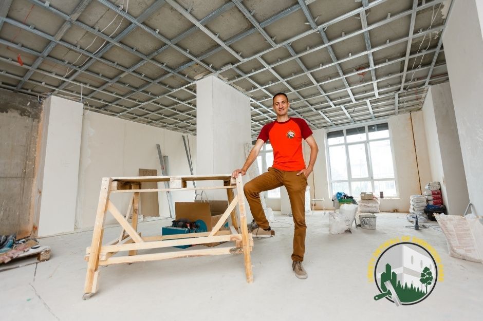 Drywall installation scene with a man standing confidently inside an unfinished residential room, one foot resting on a wooden work stand. The space shows exposed ceiling framing, tools, and construction materials scattered around Sherwood Park, Alberta.