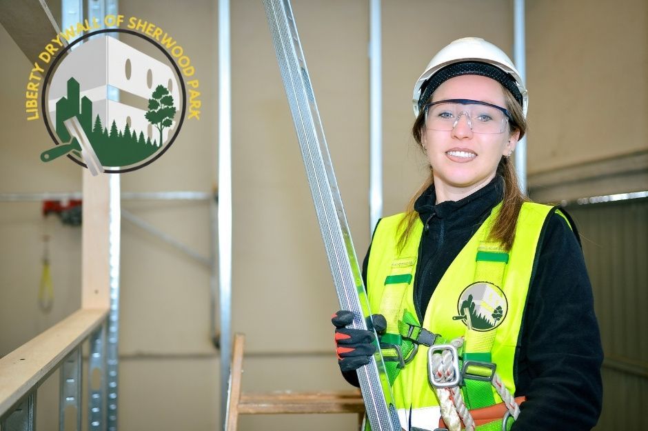 A drywall repair company worker wearing a white hard hat, safety glasses, and a yellow safety vest stands indoors holding a long metal stud used for drywall framing. She smiles confidently while standing in front of partially built residential wall frames. Located near Rivard Crescent, and Reichert Dr, Beaumont, AB.
