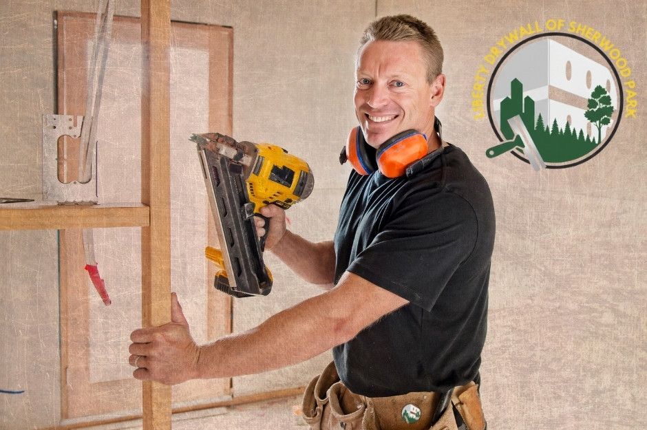 A drywall repair company worker on a commercial project holds a nail gun while installing drywall panels on wooden framing. He wears safety gear, including an orange ear protection and a tool belt near Lamplight Bay, and Lamplight Dr, Spruce Grove, AB.