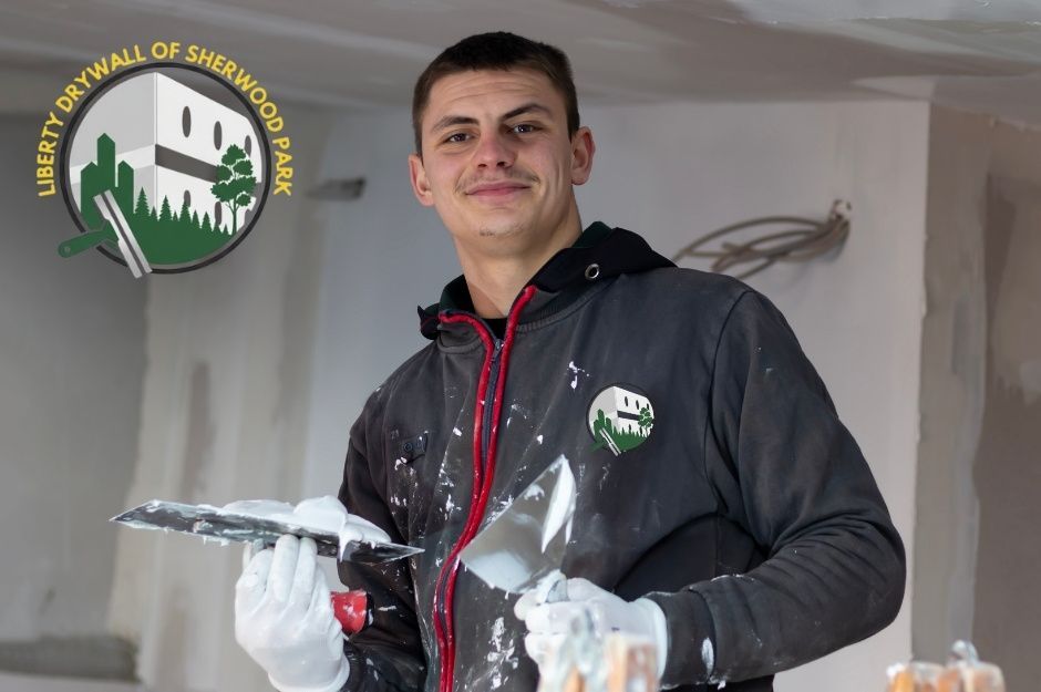 A drywall repair contractor stands indoors wearing a black hoodie with red trim and white gloves, holding a metal taping knife and a putty knife covered in joint compound, working on smoothing walls and ceilings in a commercial renovation project near Main St, and 3 Ave, Ardrossan, AB.