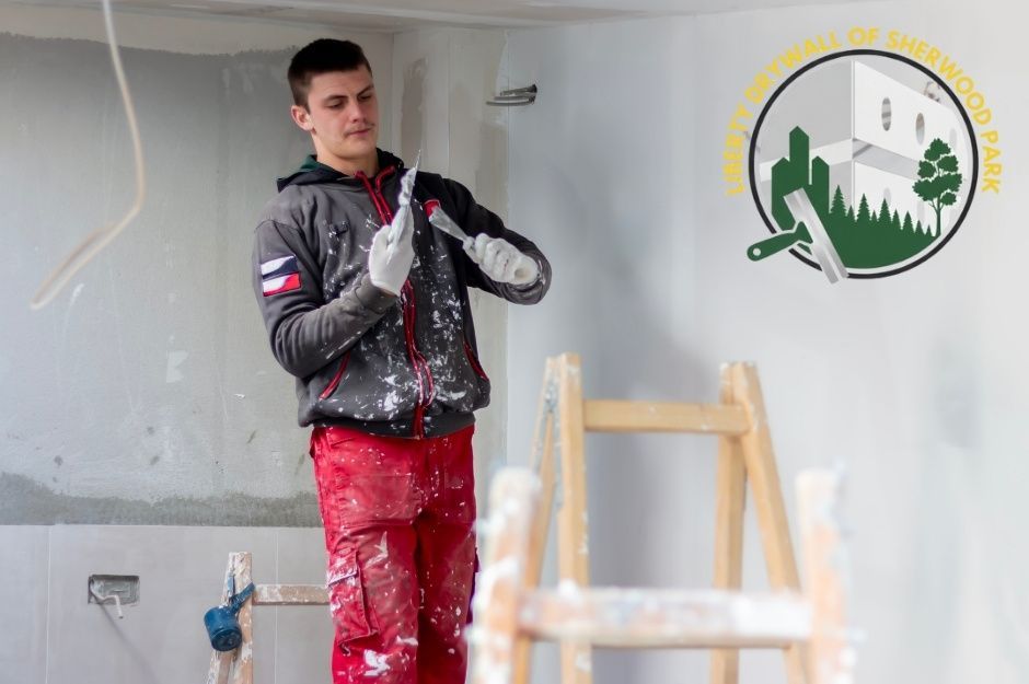 A drywall repair contractor at a commercial construction site wearing a black hoodie and red work pants, using a drywall knife to apply joint compound to the white wall, with a wooden ladder visible near 50 St, and 55 Ave, Gibbons, AB.