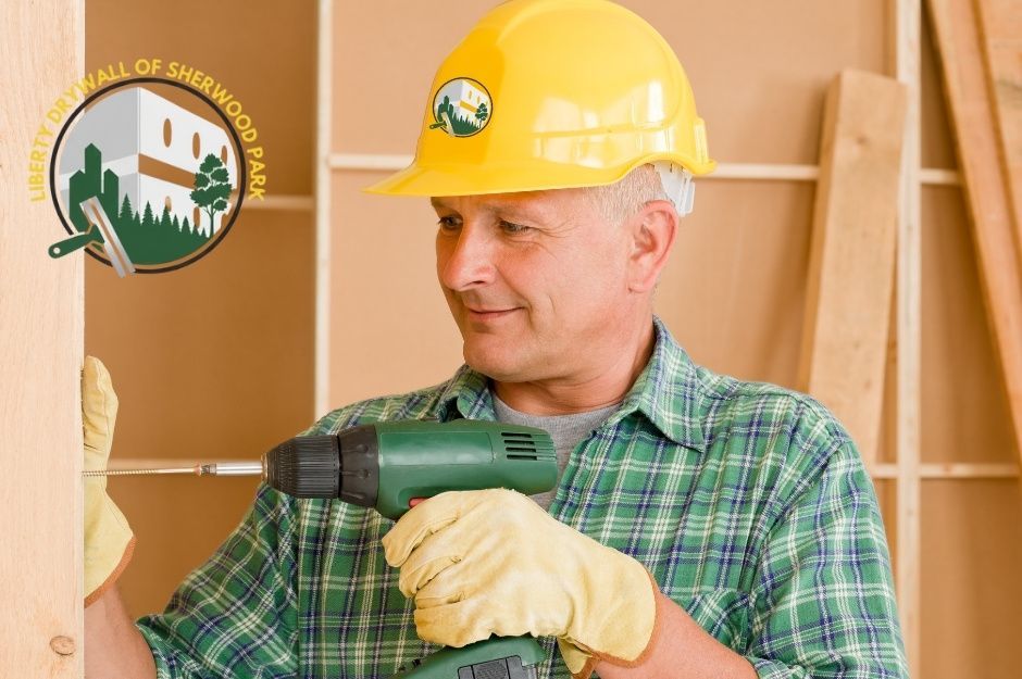 A drywall repair contractor, wearing a yellow hard hat and a green plaid shirt, works on a residential project. He is wearing work gloves and is positioned among wooden framing near Grayridge Bay, and Graybriar Dr, Stony Plain, AB.